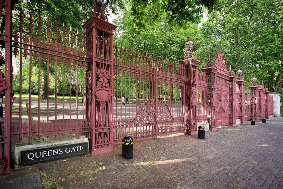 A large, decorative cast-iron gate painted in a deep reddish-pink hue, featuring ornate scrollwork and intricate detailing, stands prominently in the foreground. The gate is supported by tall, square pillars topped with decorative lantern-style streetlights, also painted in matching reddish-pink. The gate is closed and spans a wide entrance, with a small pedestrian access gate nestled within the larger structure. In the lower left corner, there is a black and white sign reading 'QUEENS GATE' positioned on the pavement. The surrounding environment includes a cobblestone driveway in front of the gate, with short black bollards featuring yellow reflective bands placed along the edge of the pavement for safety. In the background, lush green trees with dense foliage create a shaded canopy, while a glimpse of a paved pathway or street can be seen through the open sections of the gate. The scene appears well-maintained, with the ironwork showing a weathered but preserved finish, indicative of an historic or notable landmark, potentially associated with an estate or public park. The image naturally aligns with themes of private estate security, on-site clearance, or alternative waste handling, reflecting the aesthetic and functional aspects of rubbish removal or estate management services occasionally required in such settings.