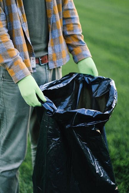 A person wearing a plaid shirt over a grey t-shirt, with a brown belt and light green gloves, is holding open a large black rubbish bag in an outdoor setting. The bag appears to be made of glossy plastic and is partially filled with waste or debris. The scene is set on a grassy area, possibly a park or garden, with a well-maintained lawn in the background. The lighting is natural, indicating daylight, and the individual seems to be engaged in waste collection or rubbish removal, which relates to alternative waste handling and private disposal services provided by Waste Collection Queen's Park. The focus is on their hands, the rubbish bag, and the surrounding greenery, emphasizing outdoor rubbish removal activities typical of independent or non-local authority waste collection efforts.