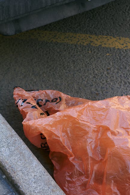 A crumpled orange plastic rubbish bag lying on an asphalt pavement beside a concrete curb, partially overlapping a dark grey textured surface that appears to be part of a road or parking area. The bag is wrinkled and semi-transparent, with black printed text partially visible on it. In the background, a vehicle's rubber tire and the lower section of a vehicle's body are visible, indicating an outdoor urban environment. The scene suggests a typical scene for waste collection or disposal outside a shop or residential area, where private or independent rubbish collection services such as those offered by waste collection companies might handle waste removal directly from the site.
