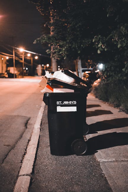 A large black wheelie bin situated on the pavement of a quiet urban street at night. The bin is labeled with the name 'St. John's' and filled with various waste items, including cardboard boxes and plastic packaging, which are visible and slightly overflowing. The bin’s wheels are on the textured asphalt, and its lid is open, revealing the contents inside. The street is dimly lit by nearby streetlights, casting soft shadows and warm tones across the scene. In the background, there are trees, buildings, and parked cars, with some illuminated windows and distant street lights creating a calm, suburban atmosphere. The scene suggests private waste collection or on-site rubbish disposal, consistent with alternative rubbish removal methods that might be handled by local waste management services like Waste Collection Queen’s Park.